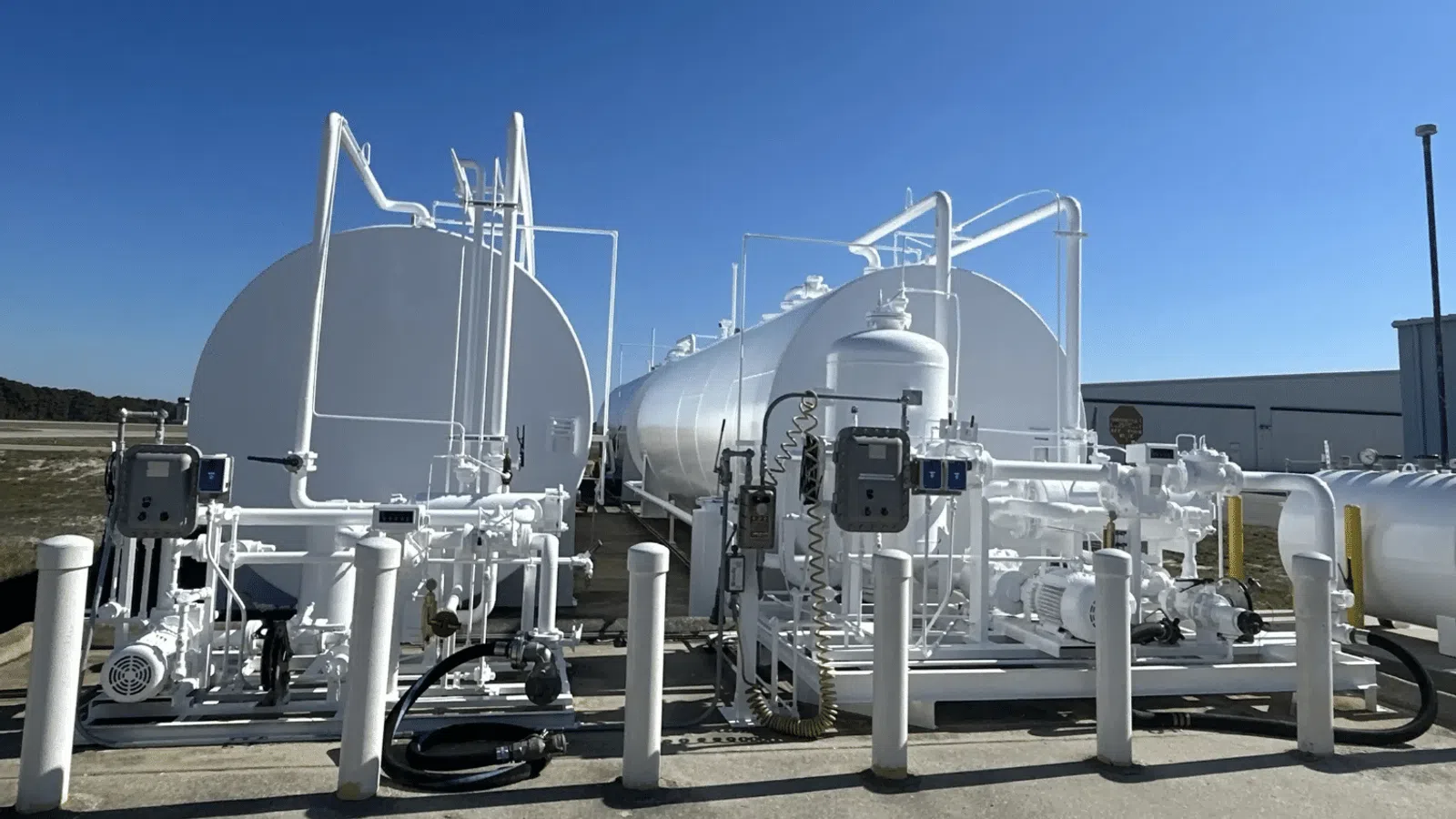 Large white industrial storage tanks with connected piping and machinery under clear blue sky outdoors.