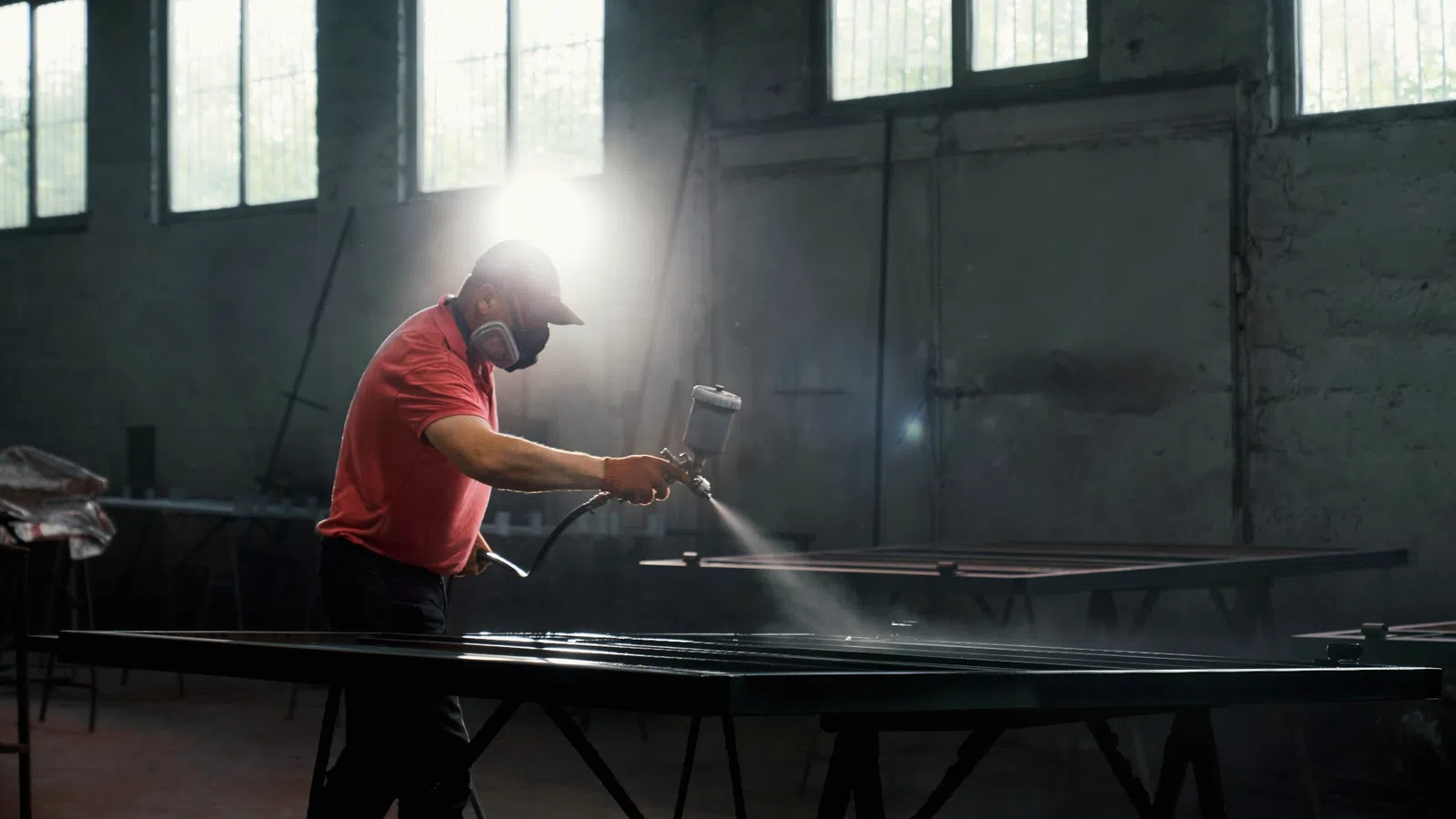 Worker wearing mask spray painting metal frames in a workshop with sunlight through windows
