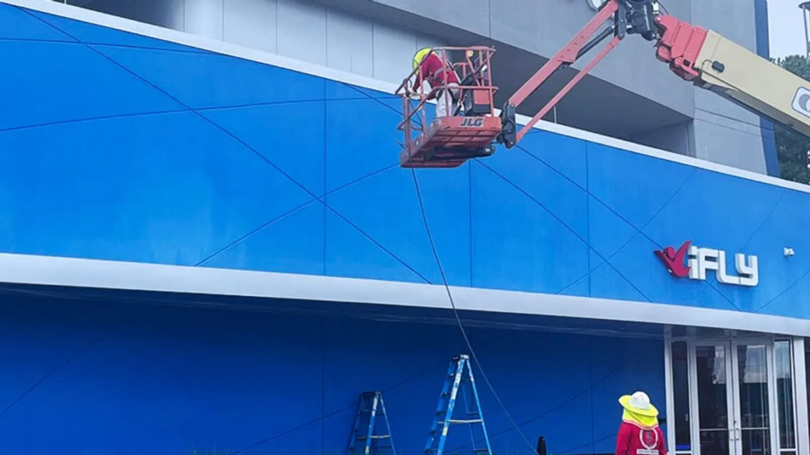 Workers use a boom lift to install signage on the blue exterior wall of an iFLY indoor skydiving center.