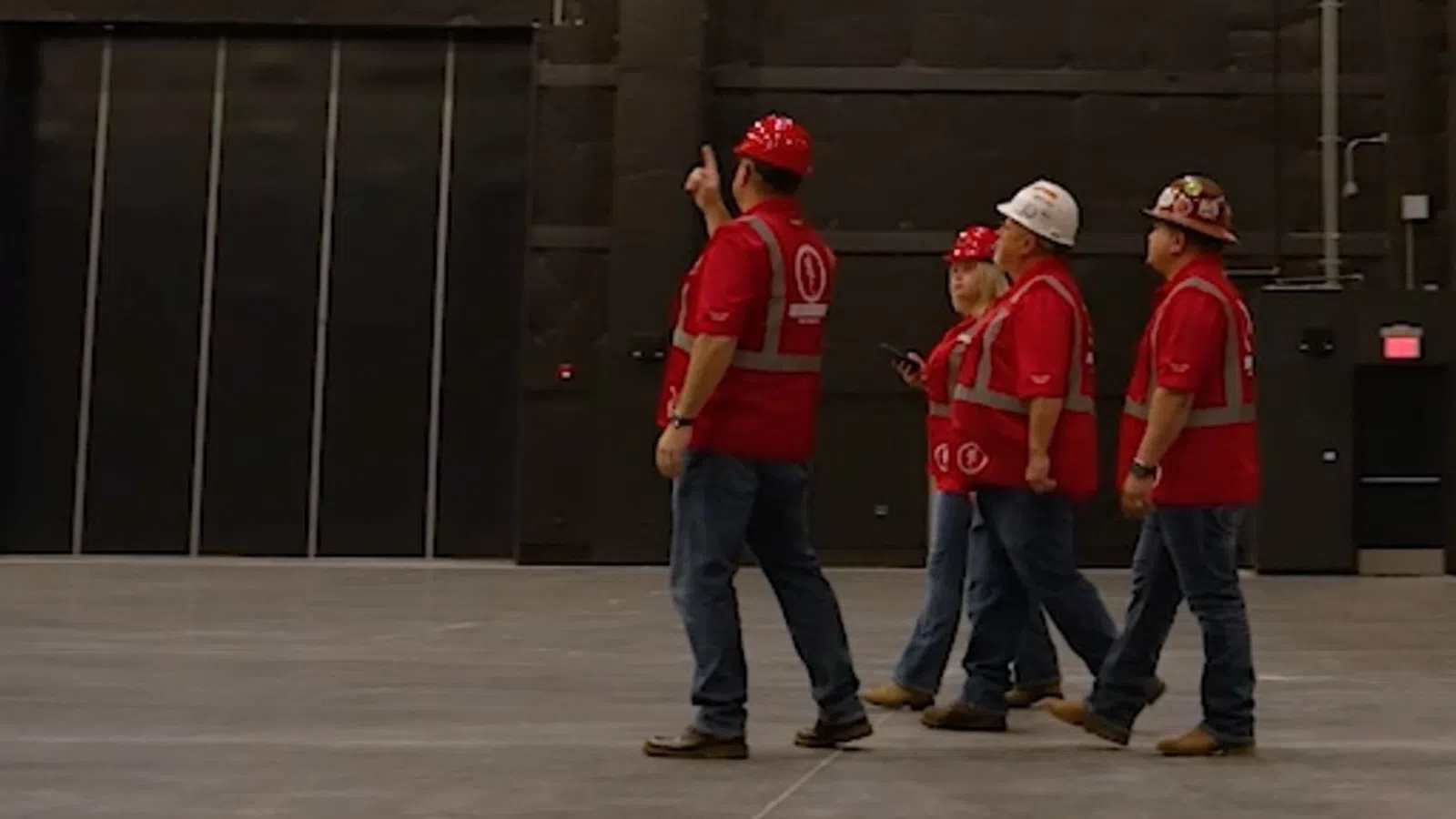 Four construction workers in red shirts and helmets walk inside a large empty warehouse or industrial space.