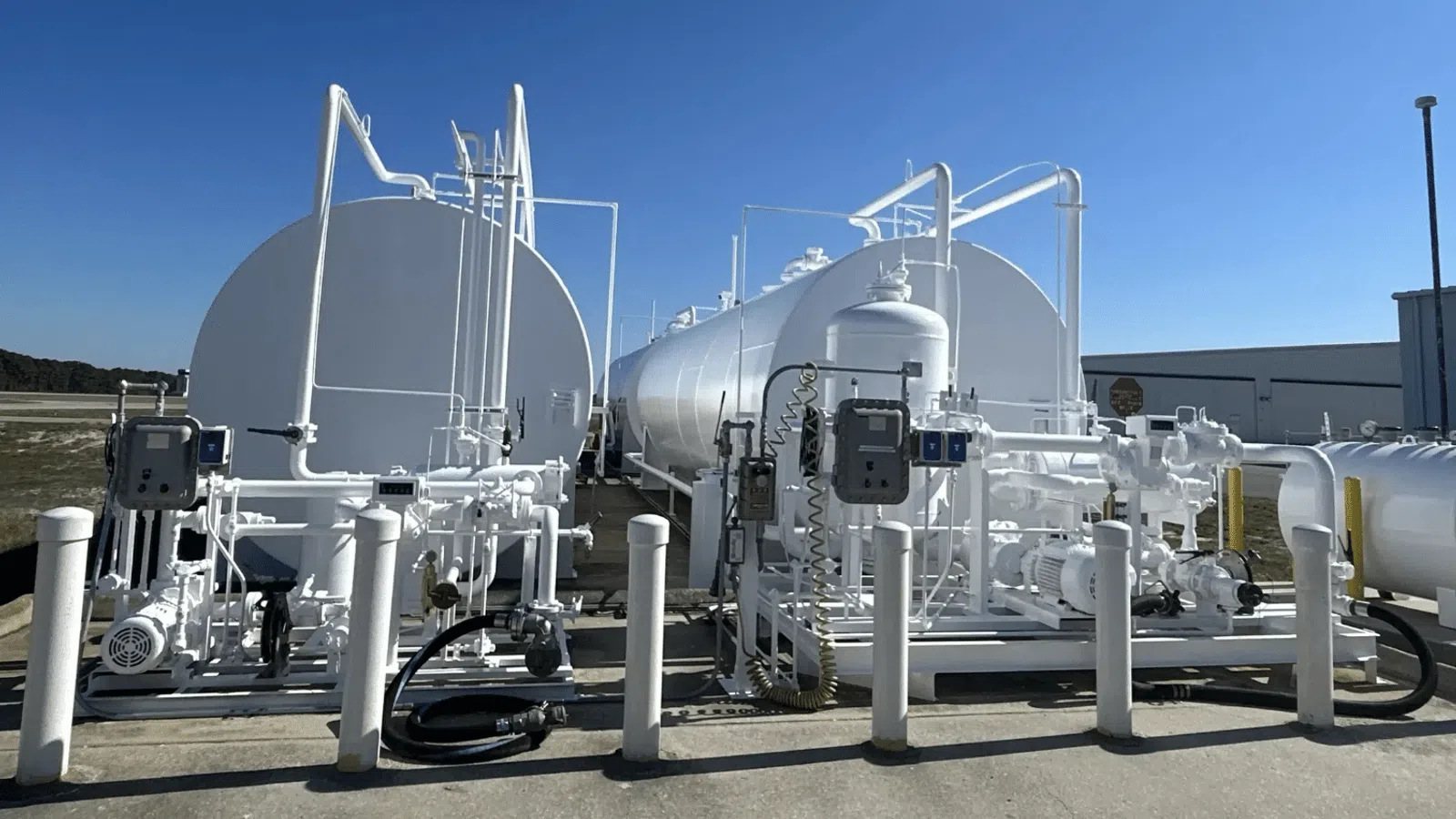 Large white industrial storage tanks with connected piping and machinery under clear blue sky outdoors.