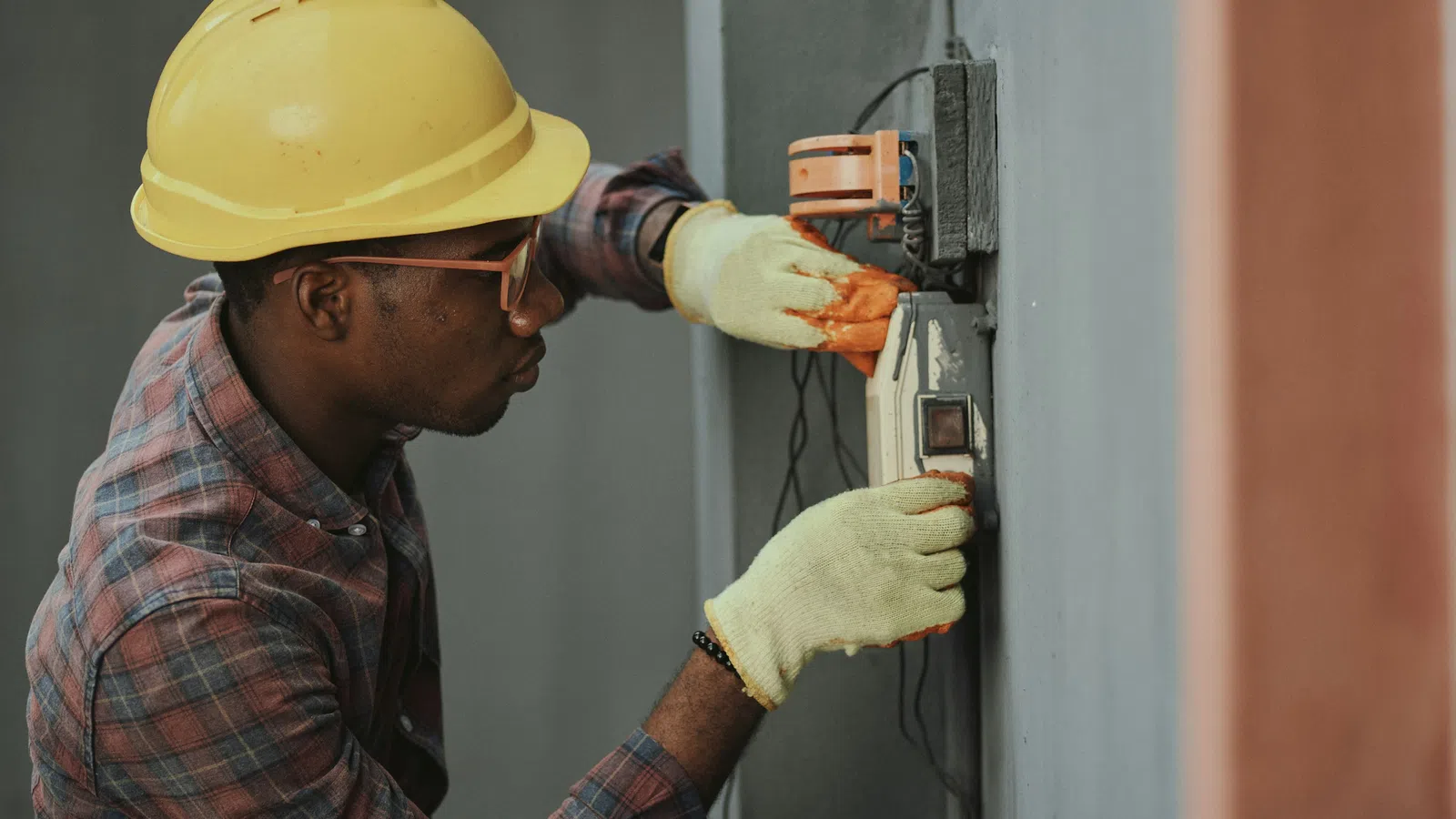 Electrician wearing hard hat and gloves working on electrical panel with tools on grey wall.