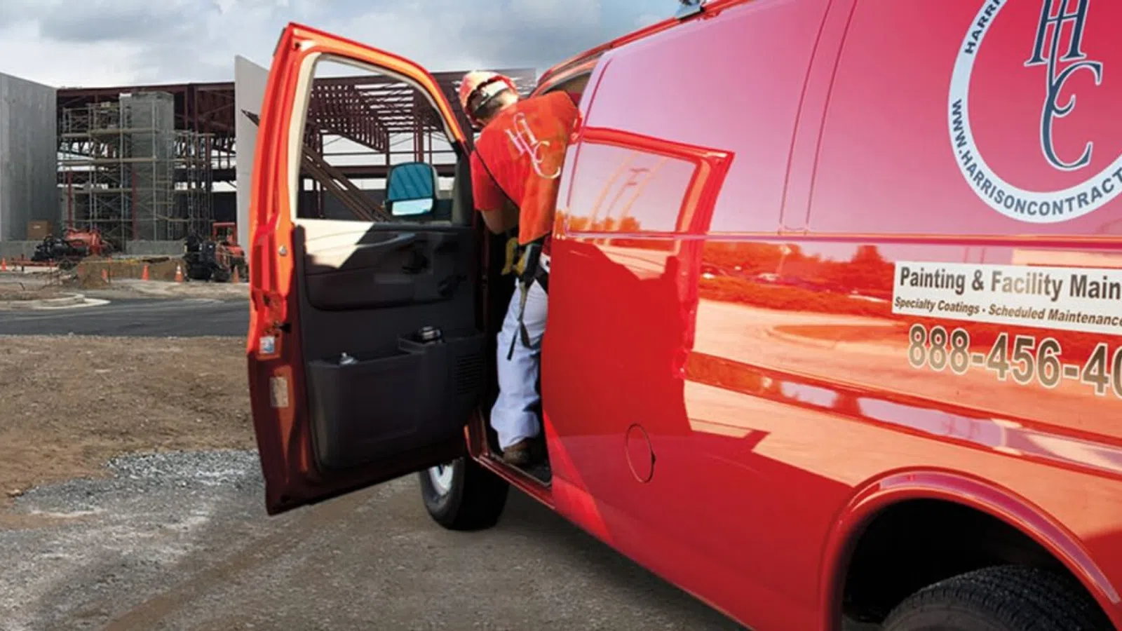 Red maintenance van with open door near construction site with steel framework under cloudy sky.