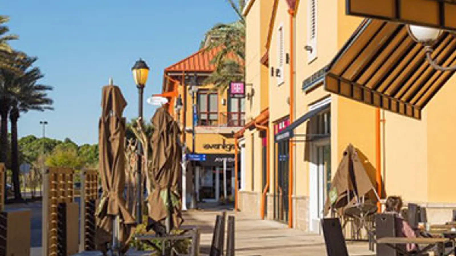 Sunny outdoor shopping plaza with yellow buildings, closed umbrellas, and empty patio tables under clear blue sky