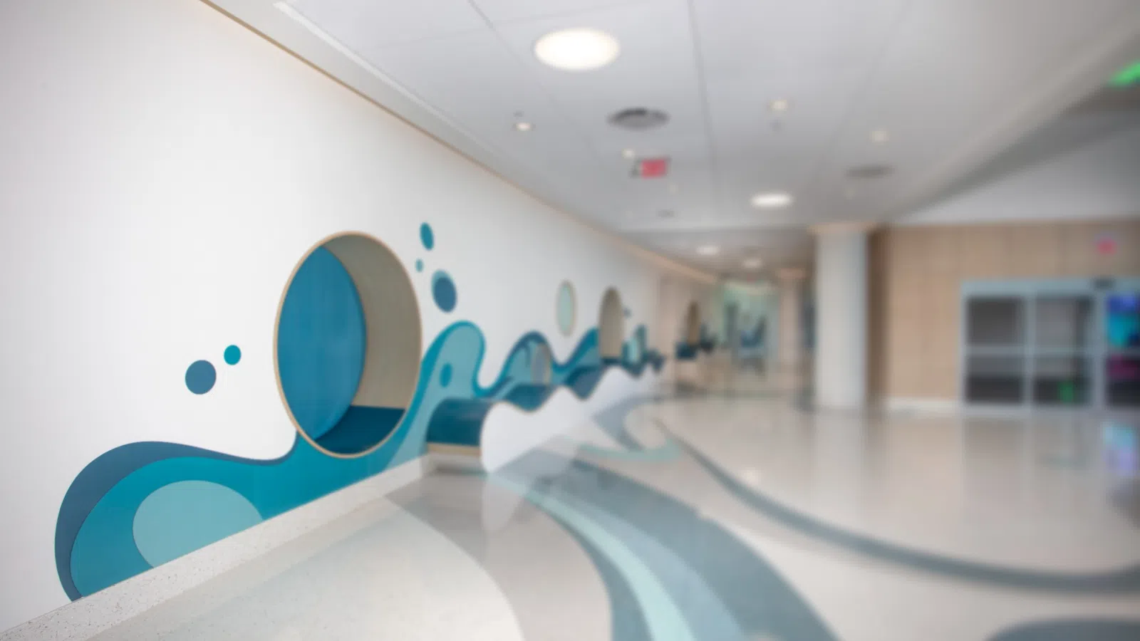 Modern hallway featuring blue wave mural with circular cutouts and smooth curved floor patterns in bright space.