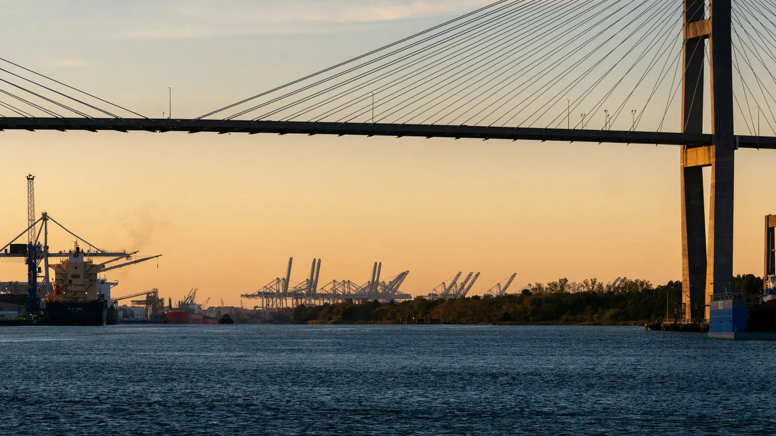 Cable-stayed bridge over river at sunset with cargo ships and industrial cranes in the background.