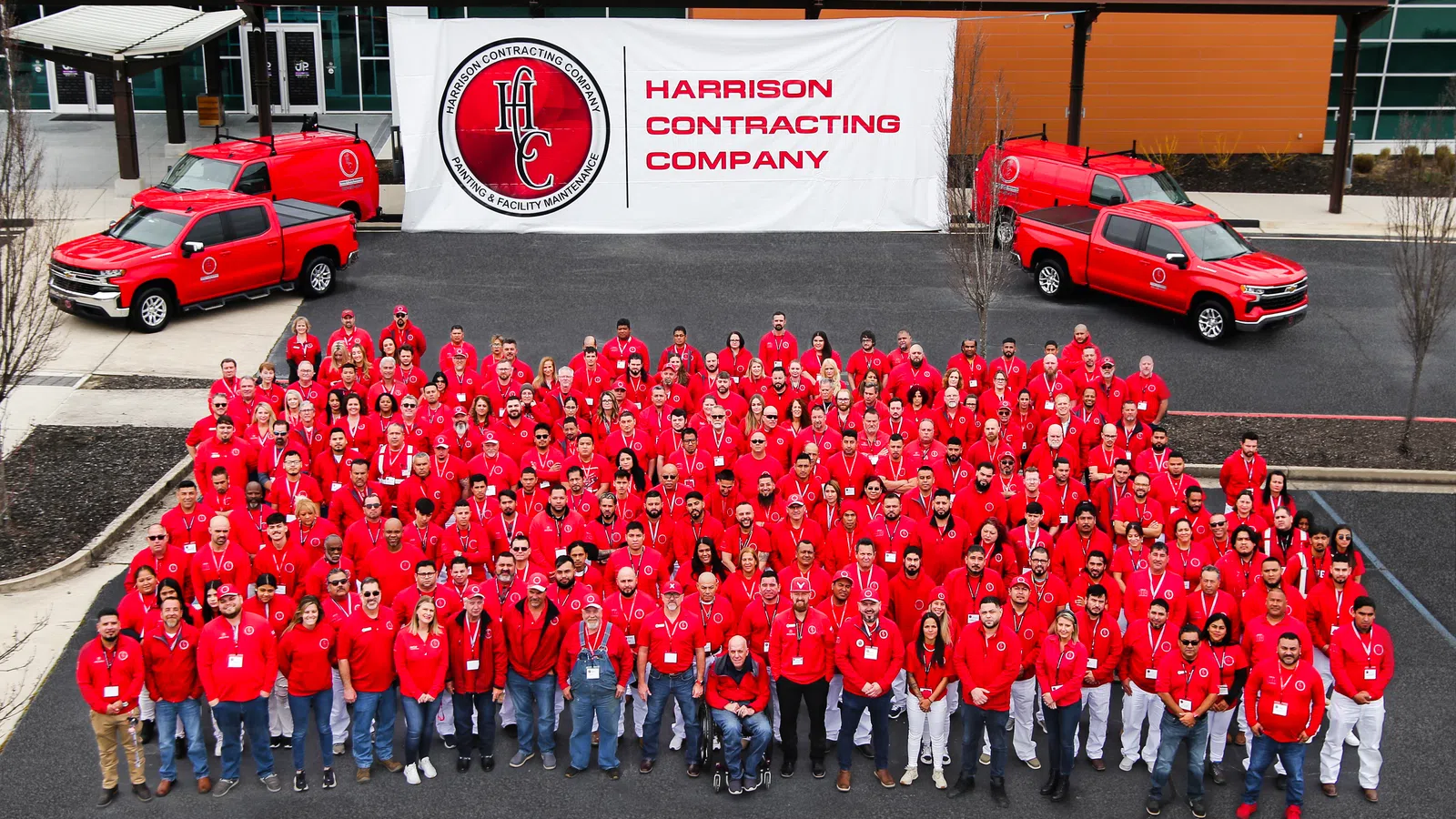 Large group of Harrison Contracting Company employees in red shirts posing outdoors with company trucks and banner
