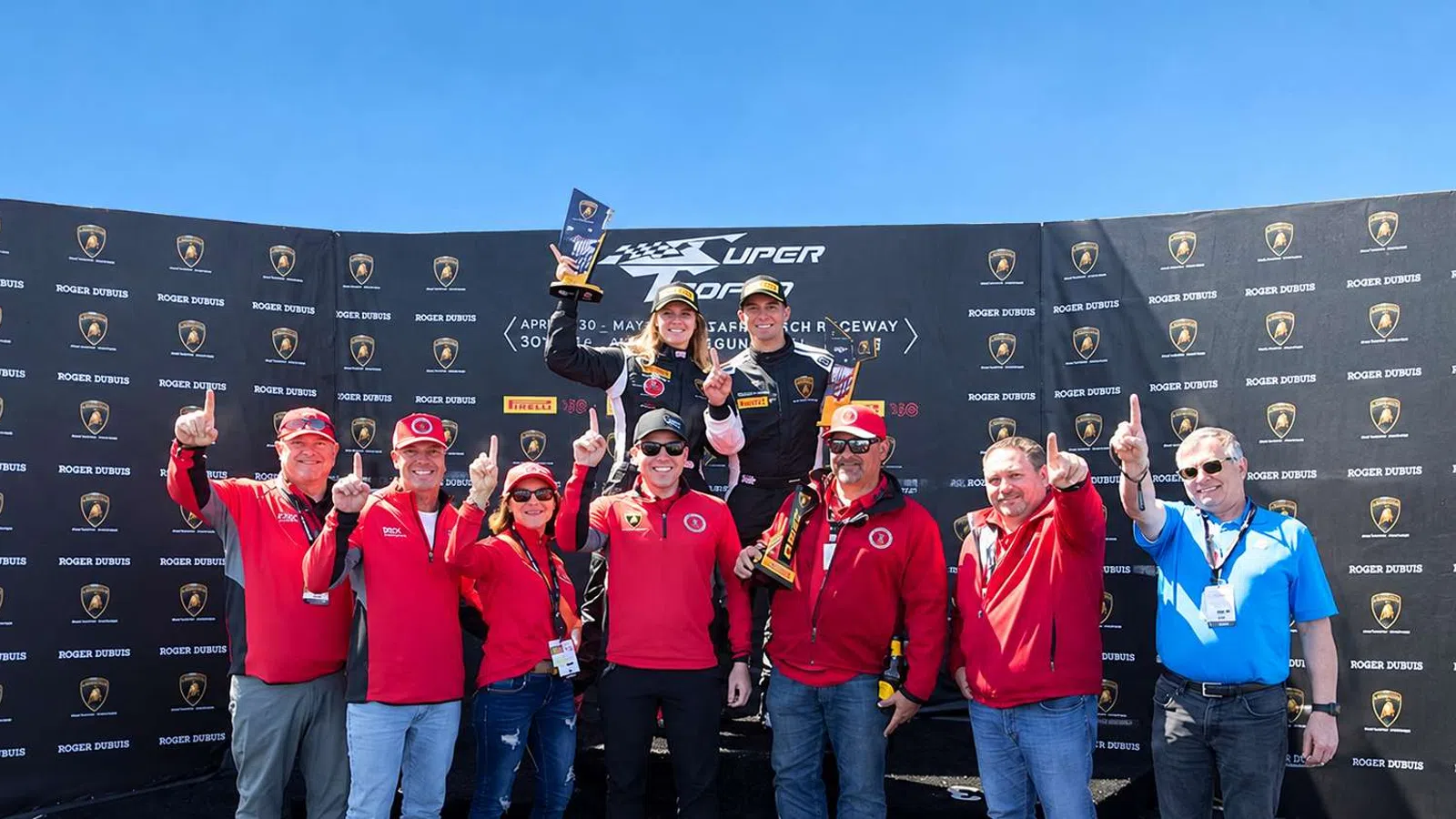 Racing team celebrating on podium holding trophies with Lamborghini and Pirelli banners in background