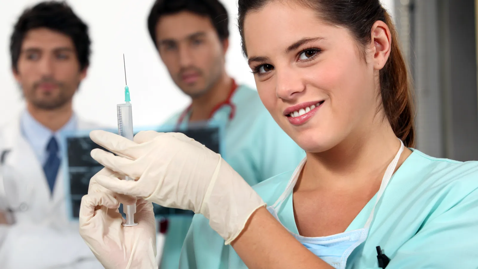Smiling nurse wearing gloves prepares a syringe with two male doctors observing behind her.