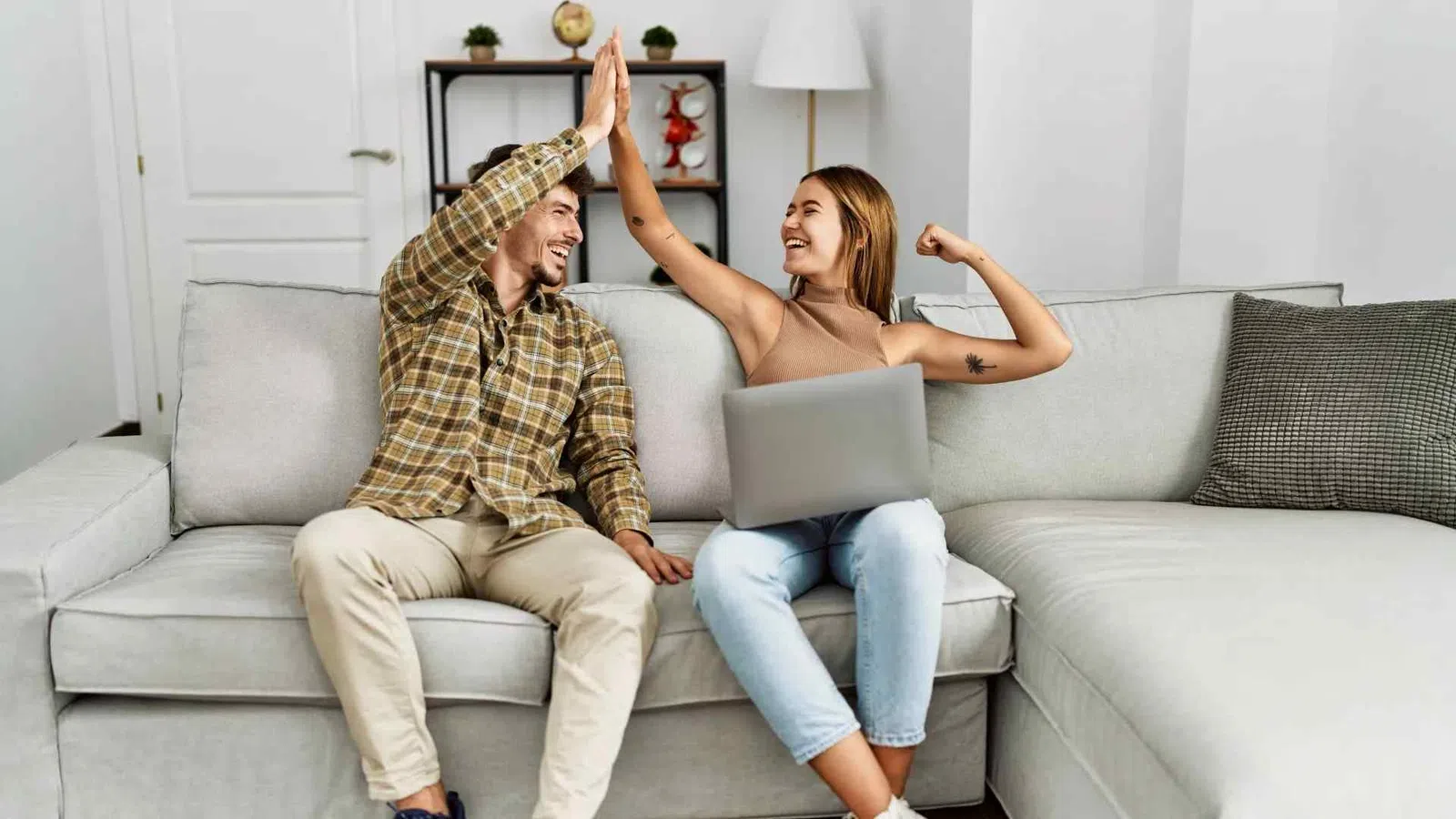 Young man and woman sitting on couch giving high five with laptop, celebrating success or achievement at home