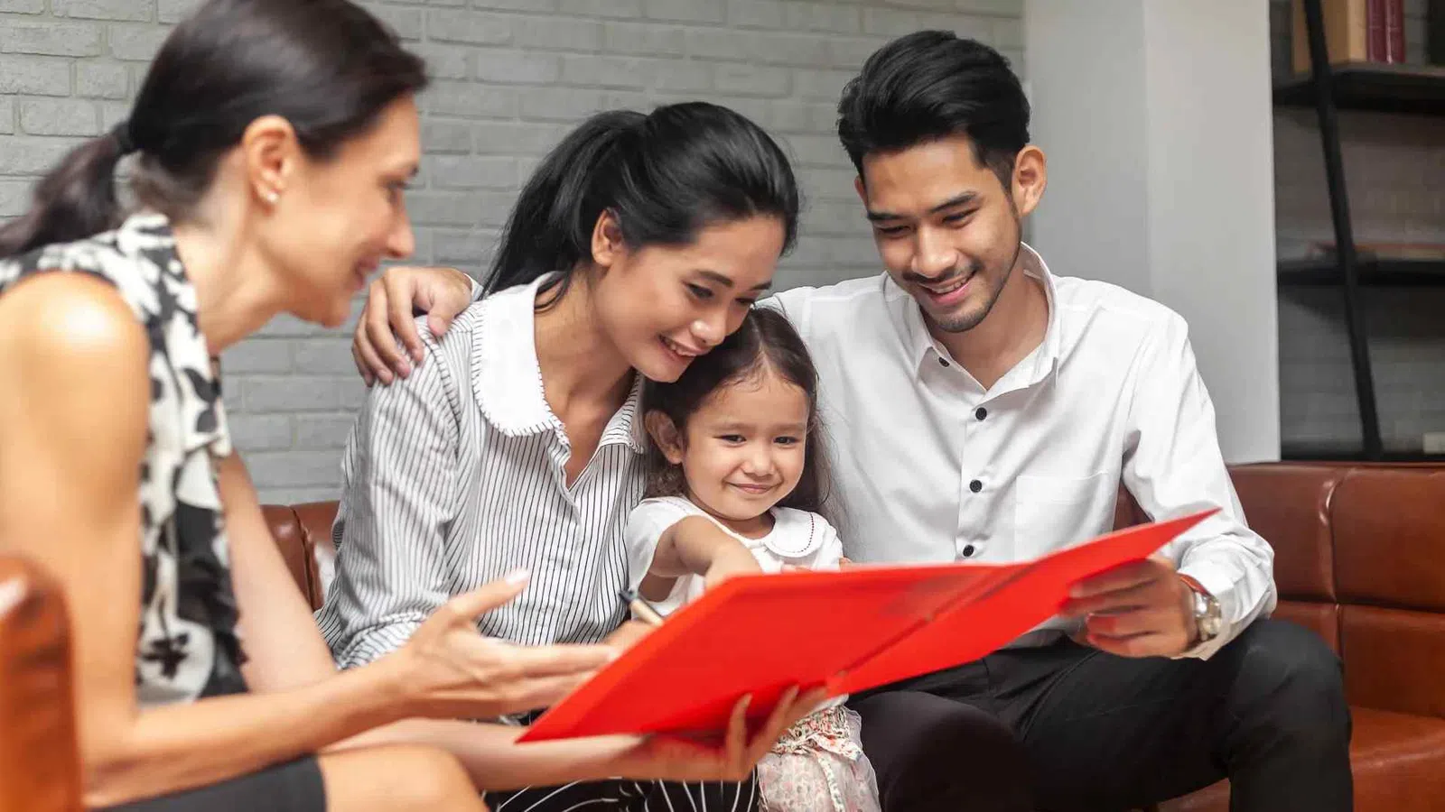 Family with young daughter smiling while looking at a red photo album in a cozy living room.