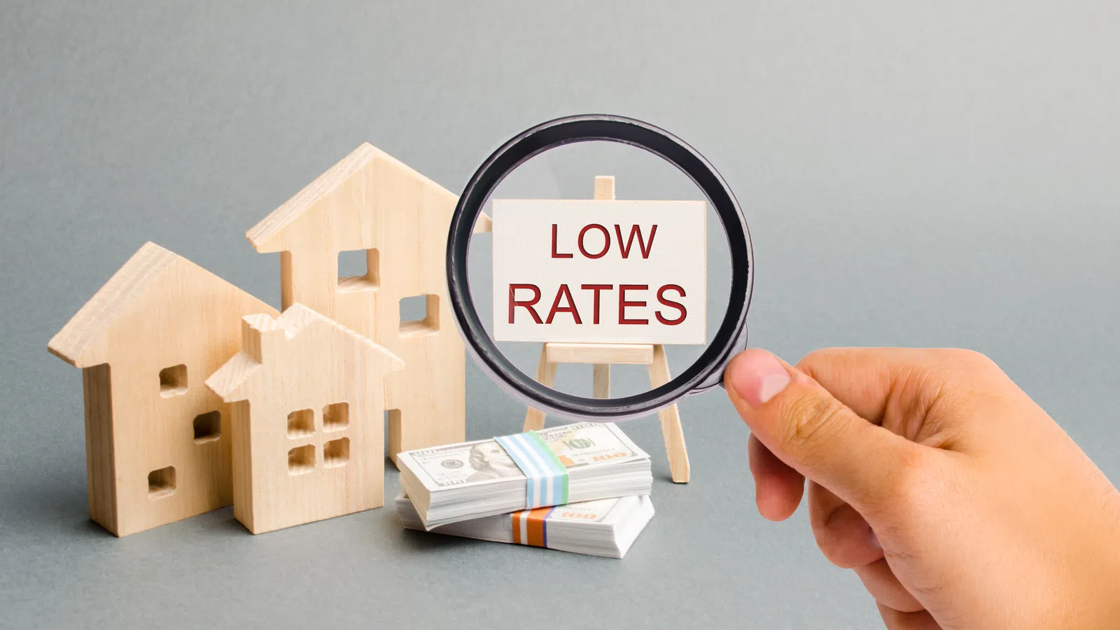 Hand holding magnifying glass focusing on low rates sign near wooden houses and money stacks