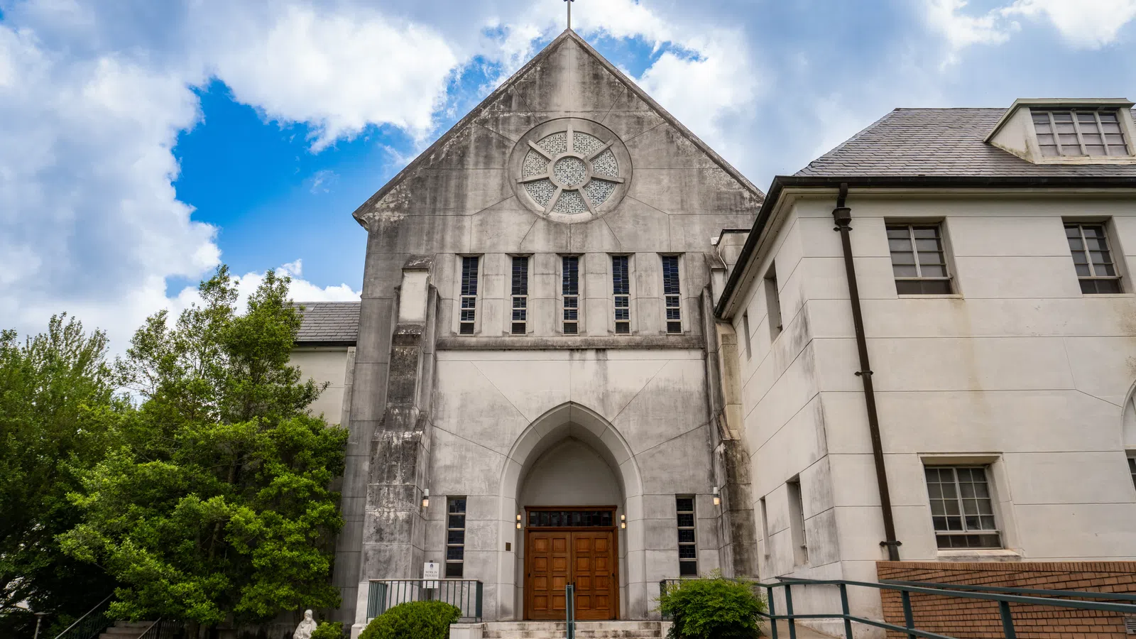 Exterior view of a historic stone church with a wooden door, cross, and trees under a blue sky with clouds