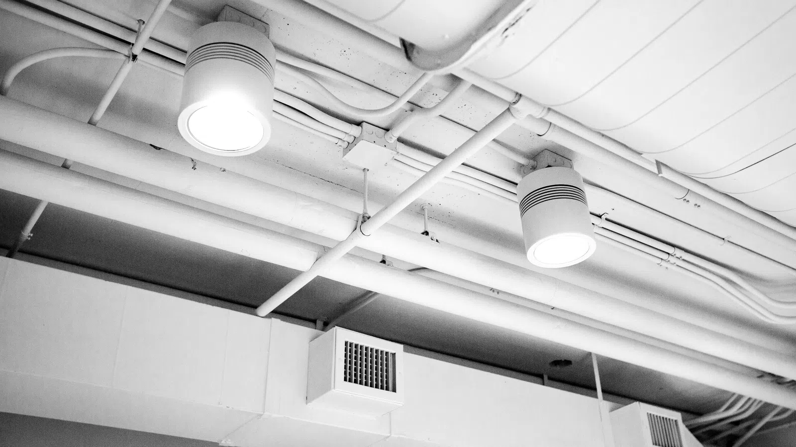 Ceiling with exposed white pipes, air vents, and two round ceiling lights illuminated in a monochrome setting