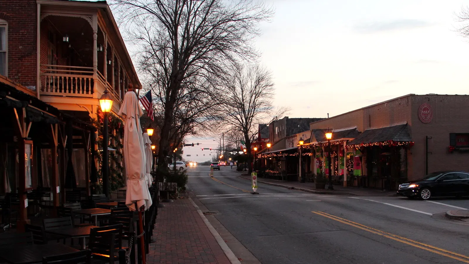 Empty street scene at dusk with historic buildings, street lamps, and outdoor seating on a small town main street.