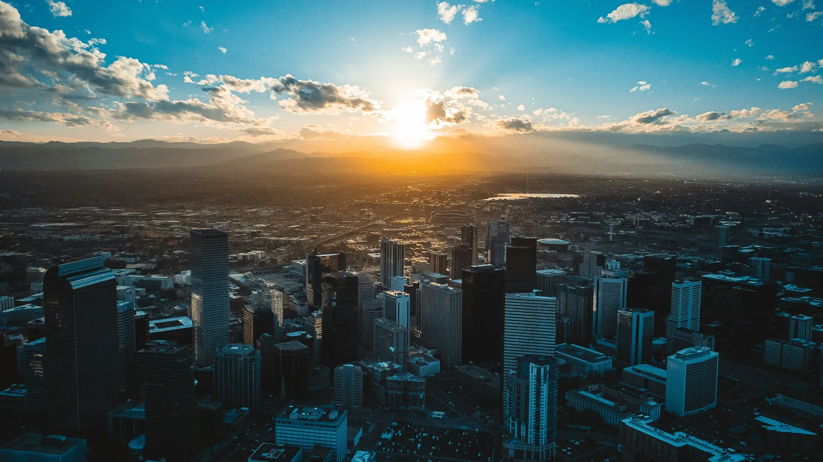 Denver skyline at sunset with sun setting behind distant mountains and scattered clouds in the sky.