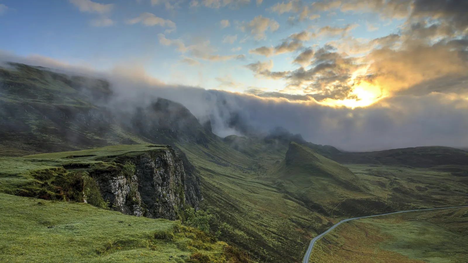 Sunset over misty cliffs and winding road in a green mountainous landscape with partly cloudy sky.