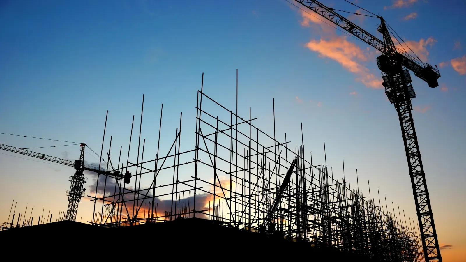 Construction site scaffolding and cranes silhouetted against a colorful sunset sky with clouds.