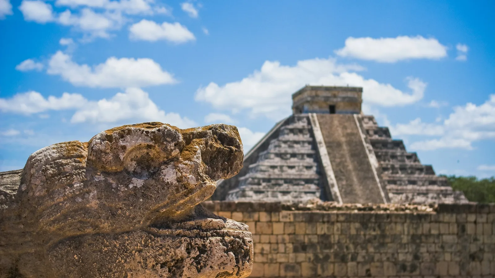 Stone serpent sculpture in foreground with ancient Mayan pyramid of Chichen Itza under blue sky.