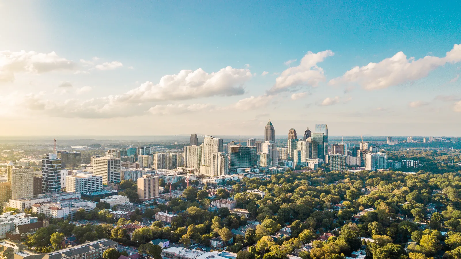 Aerial view of a city skyline with modern skyscrapers and lush green trees under a blue sky with soft clouds.
