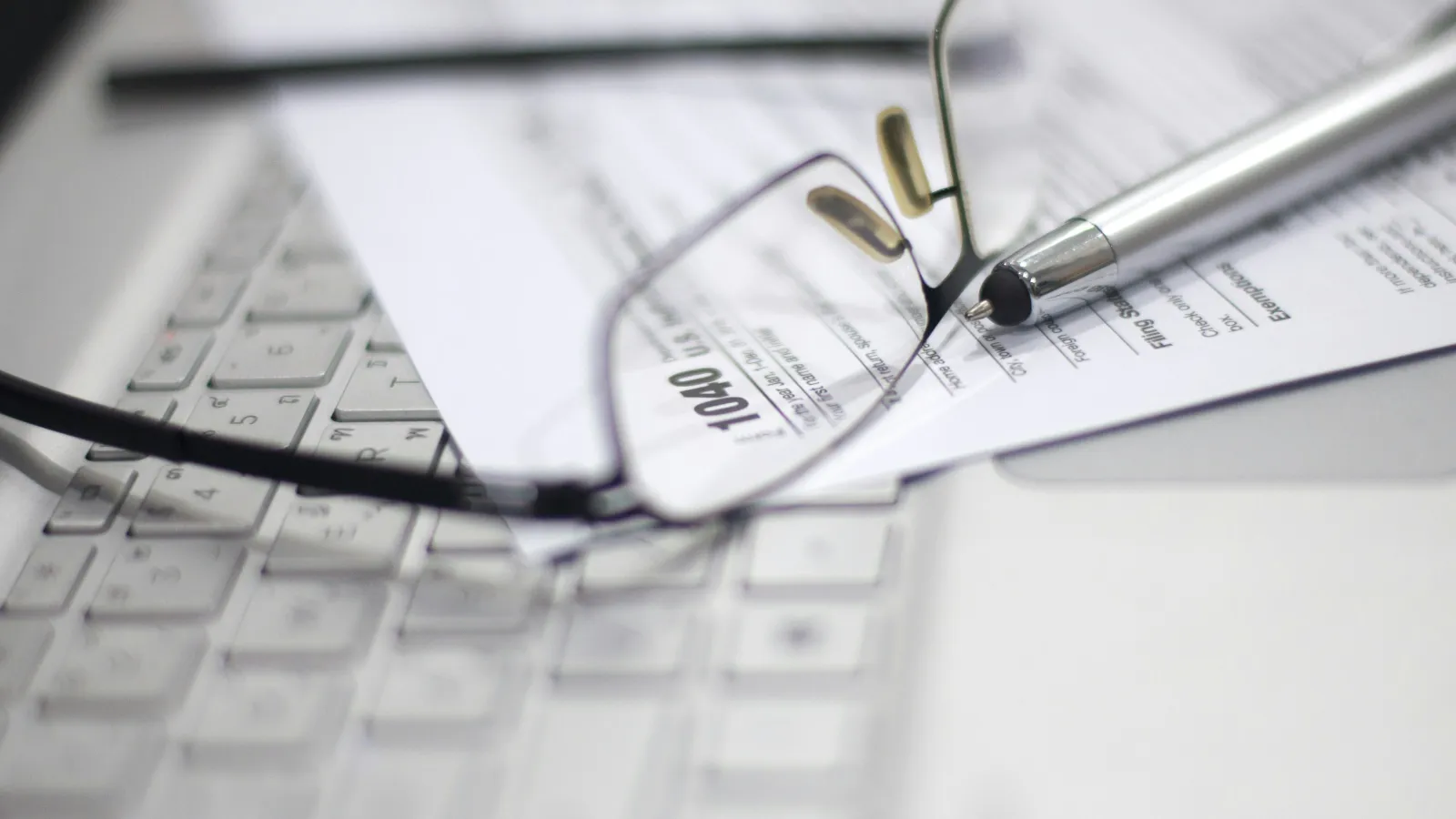 Eyeglasses and pen resting on tax form 1040 over a white keyboard, symbolizing tax preparation and filing.