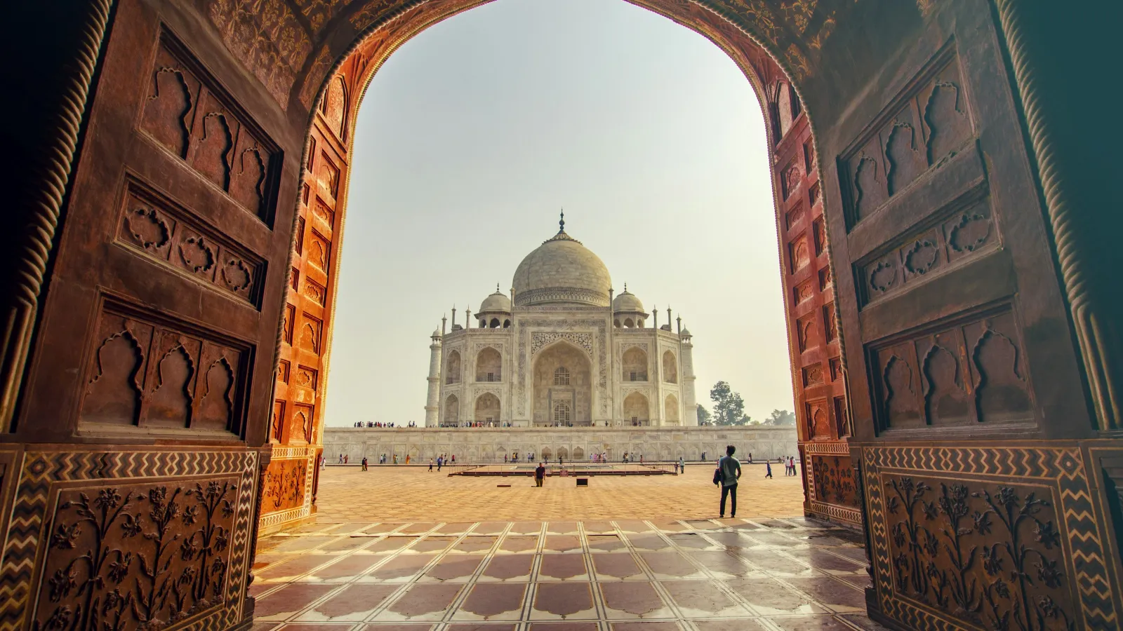 View of the Taj Mahal framed by ornate red sandstone archway with visitors in the courtyard on a clear day
