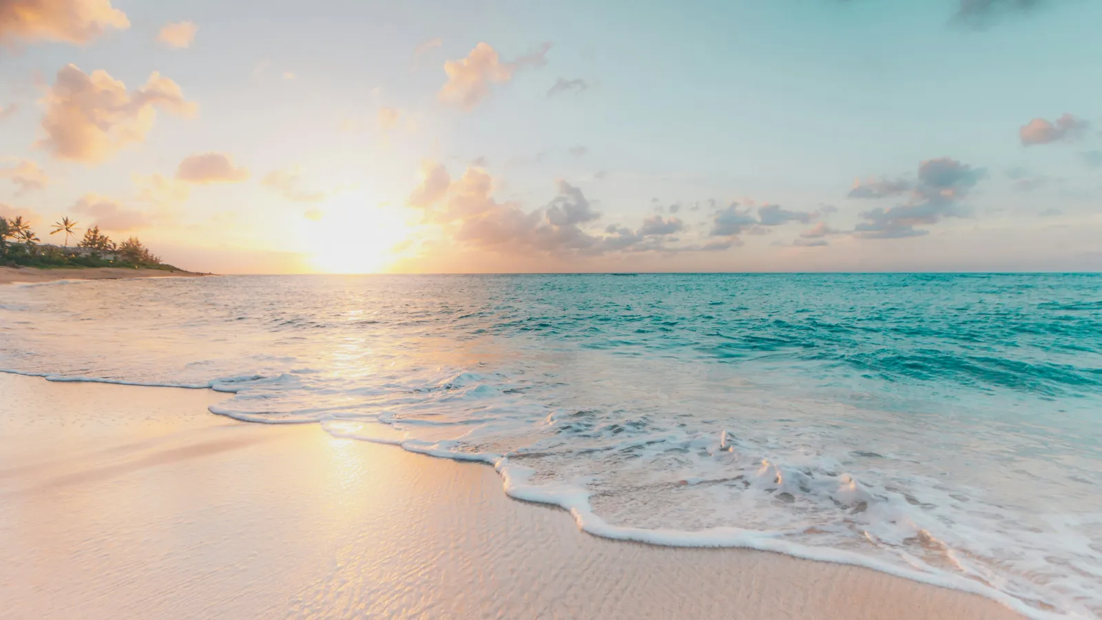 Sunset over a serene beach with gentle waves, soft sand, and a partly cloudy sky with palm trees.