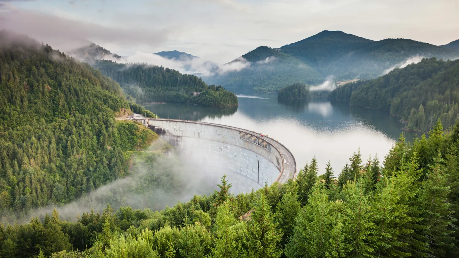 Curved dam surrounded by dense green forest and mist, with calm lake and mountains in the background at sunrise.