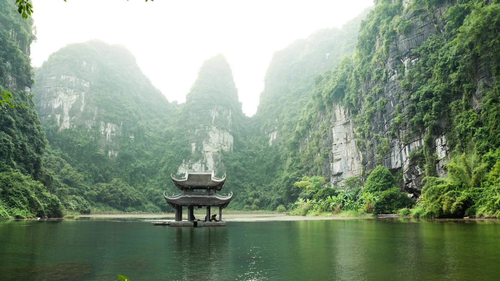 Traditional pavilion on serene lake surrounded by lush green cliffs and misty mountains in the background