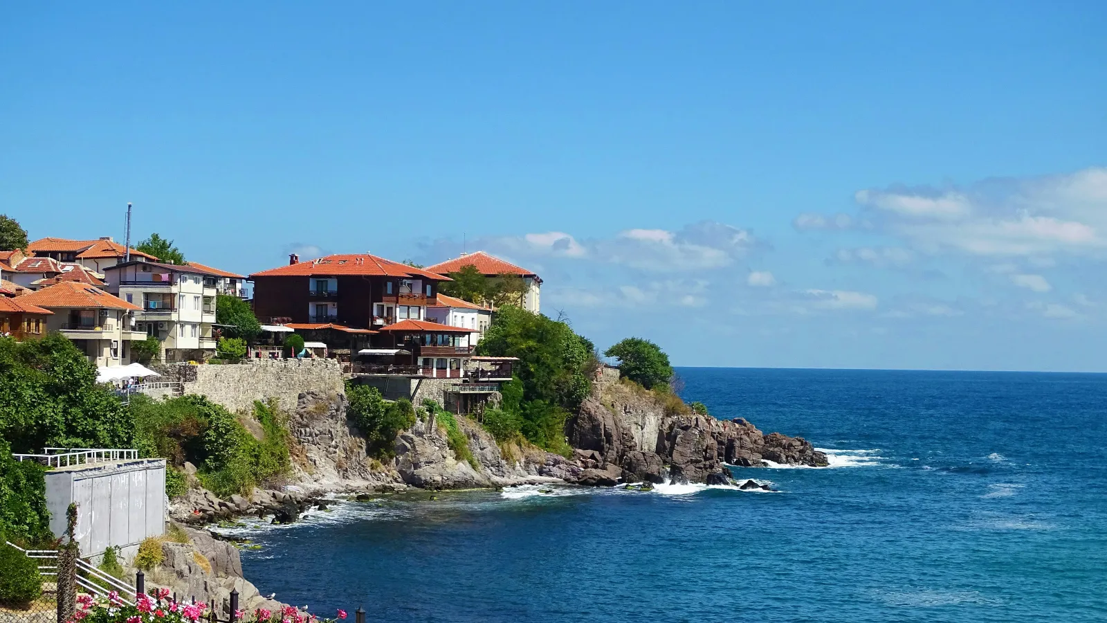 Coastal village with red-roof houses on rocky cliffs overlooking the blue sea and pink flowers in foreground.