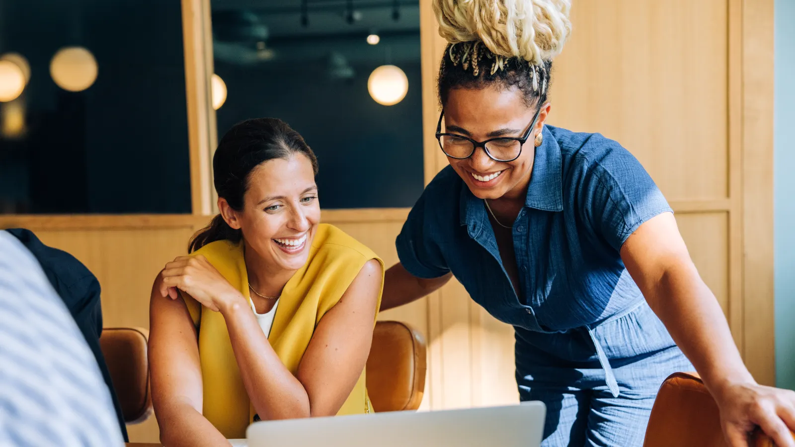 Two women smiling and collaborating while looking at a laptop in a modern office setting
