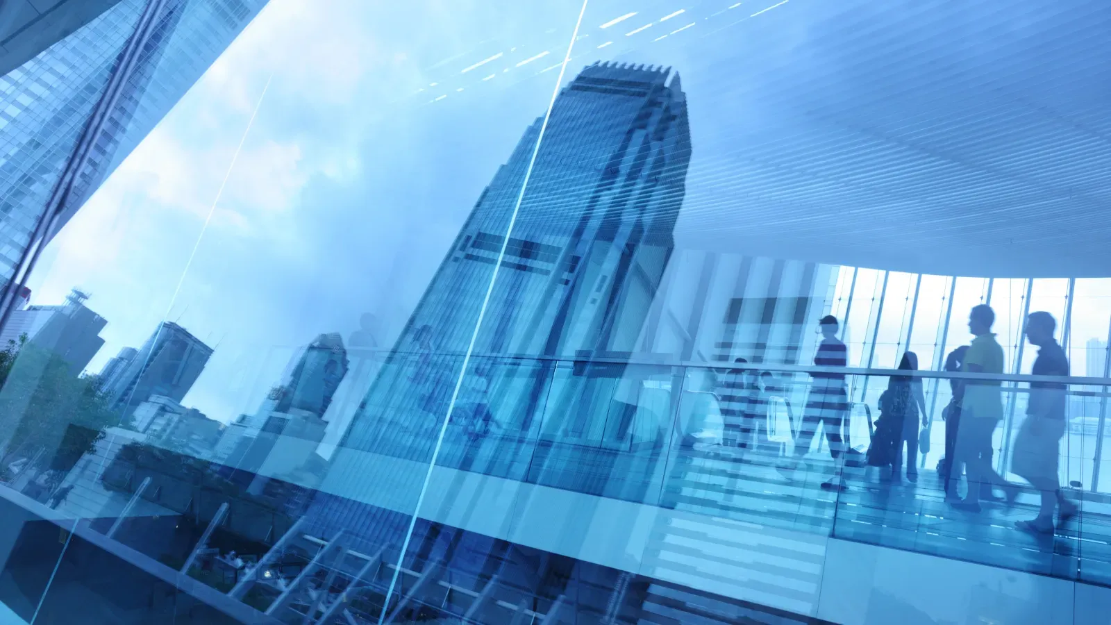 Silhouettes of people walking on a modern glass walkway with a city skyline and tall skyscraper in background.