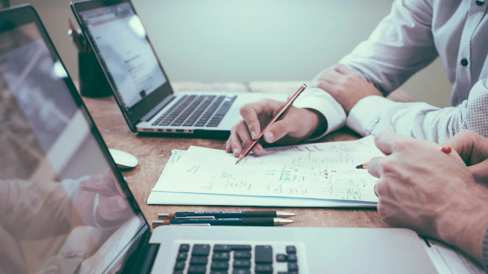 Two people working on laptops and reviewing handwritten notes on a document at a wooden desk.
