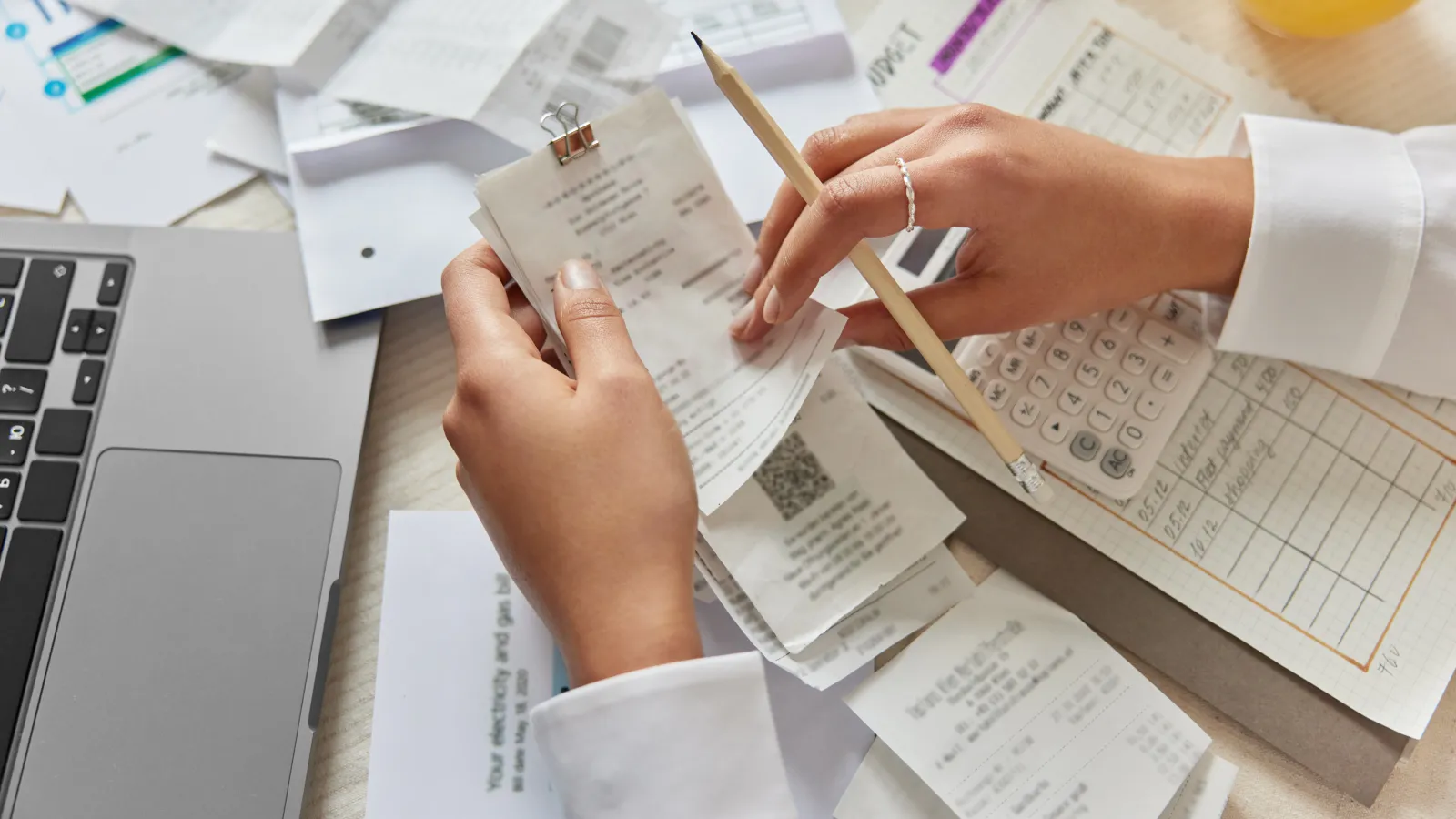 Person organizing receipts with pencil and calculator on a cluttered desk with laptop and documents.