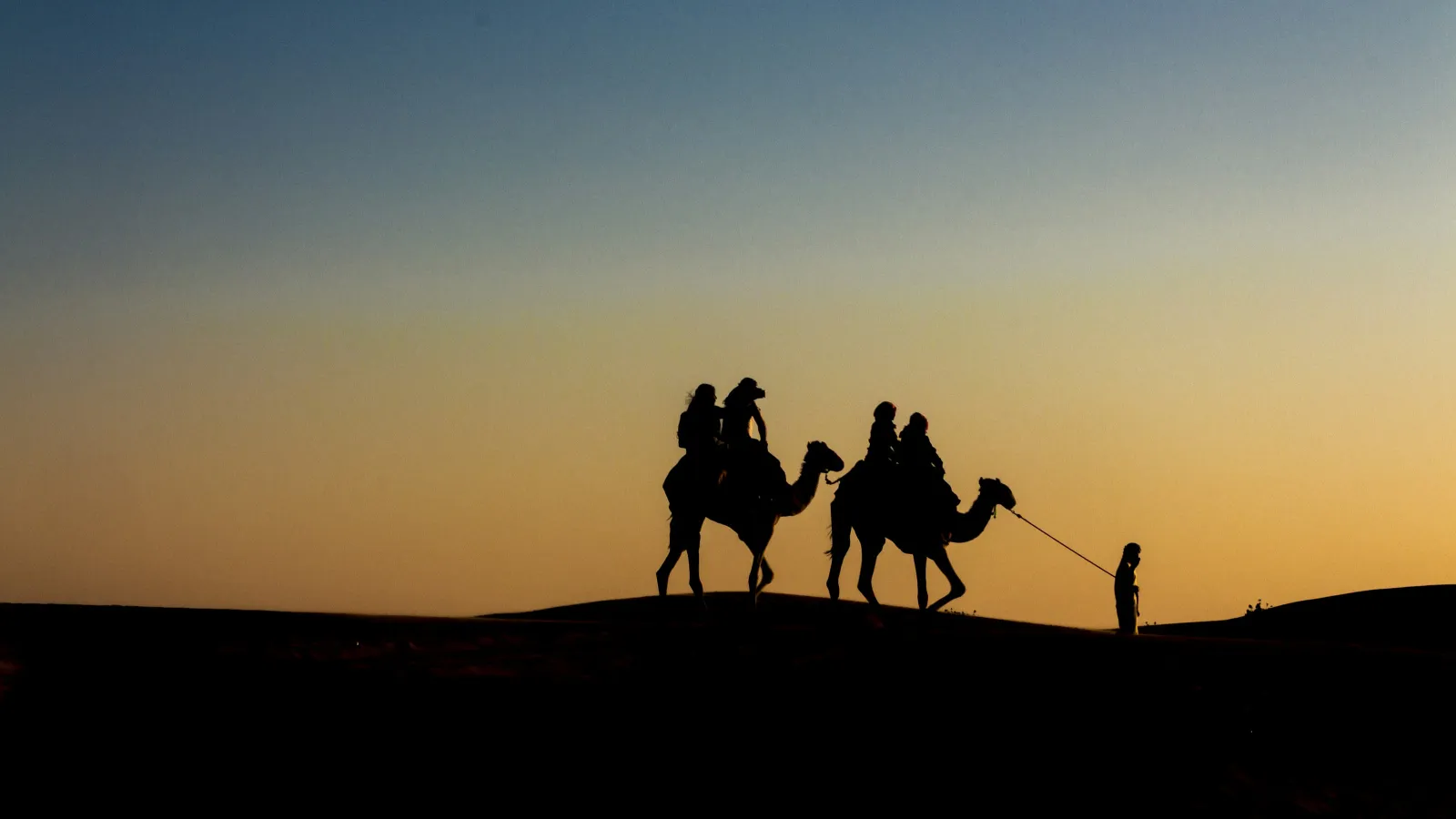 Silhouettes of people riding camels led by a person walking at sunset over desert dunes