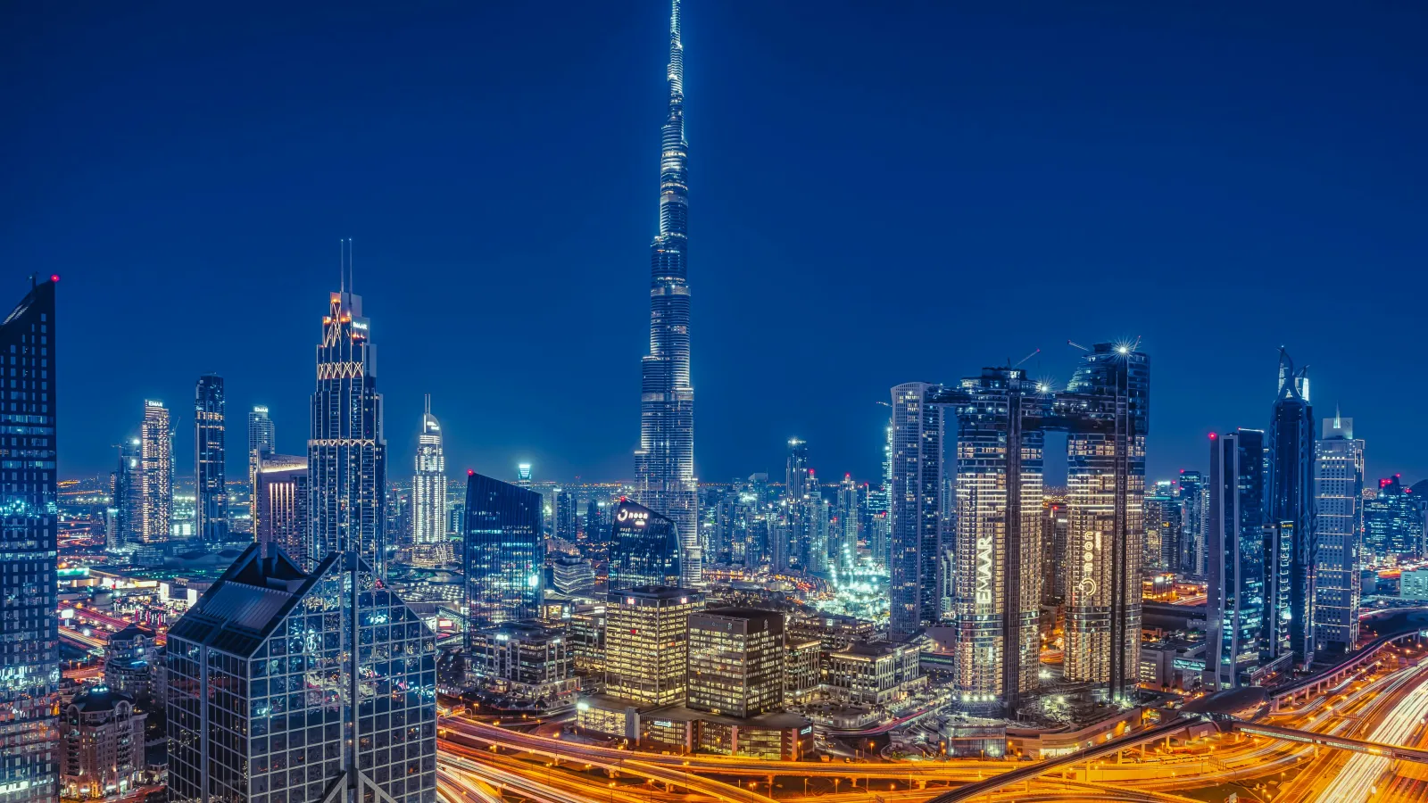 Dubai skyline at night showing illuminated skyscrapers including Burj Khalifa and busy highways with light trails.