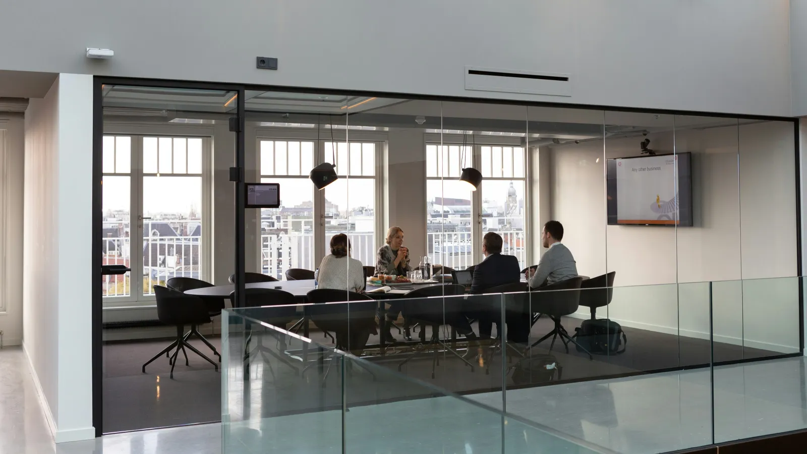 Four colleagues having a business meeting in a modern glass-walled conference room with city views outside.