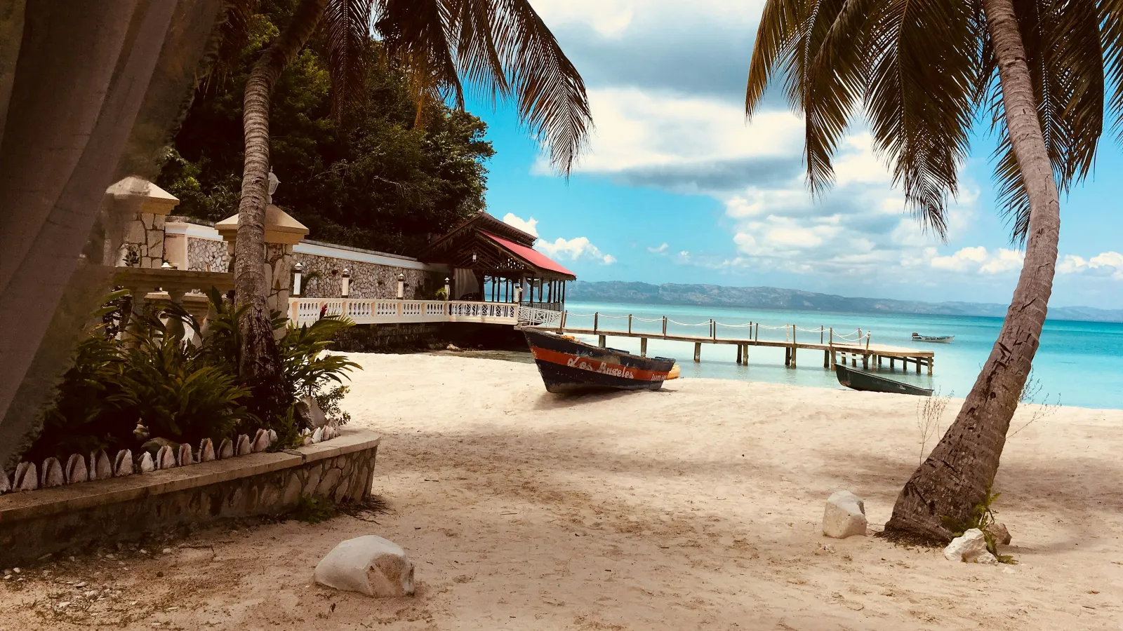 Tropical beach scene with palm trees, sandy shore, wooden pier, and boats near clear blue water under partly cloudy sky