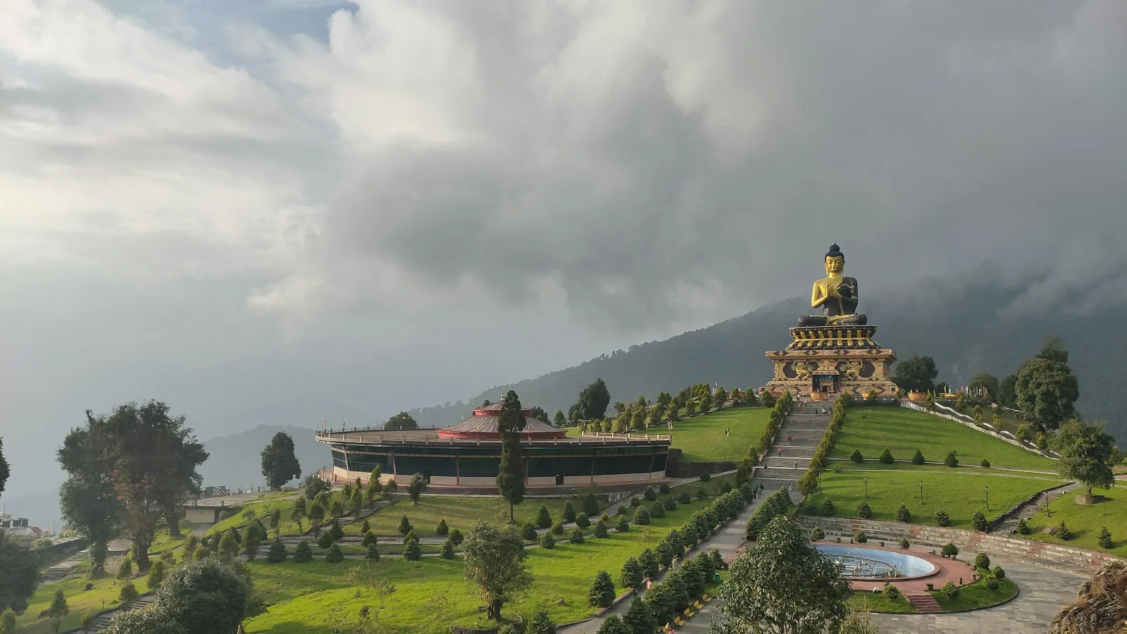 Large Buddha statue atop a hill surrounded by green gardens, pathways, and cloudy sky in a mountainous area.