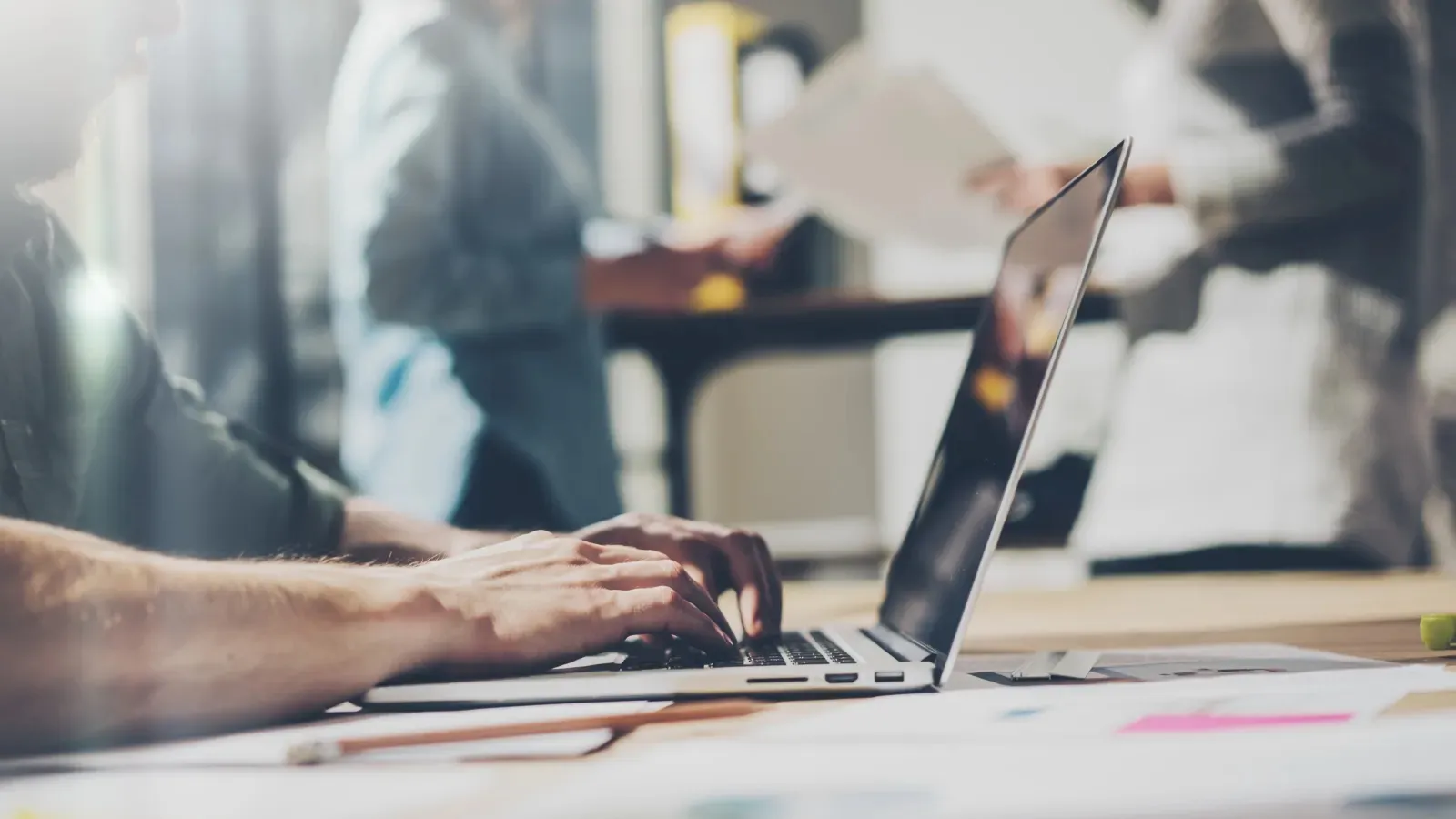 Person typing on a laptop keyboard with blurred people and documents in the background in a modern office.
