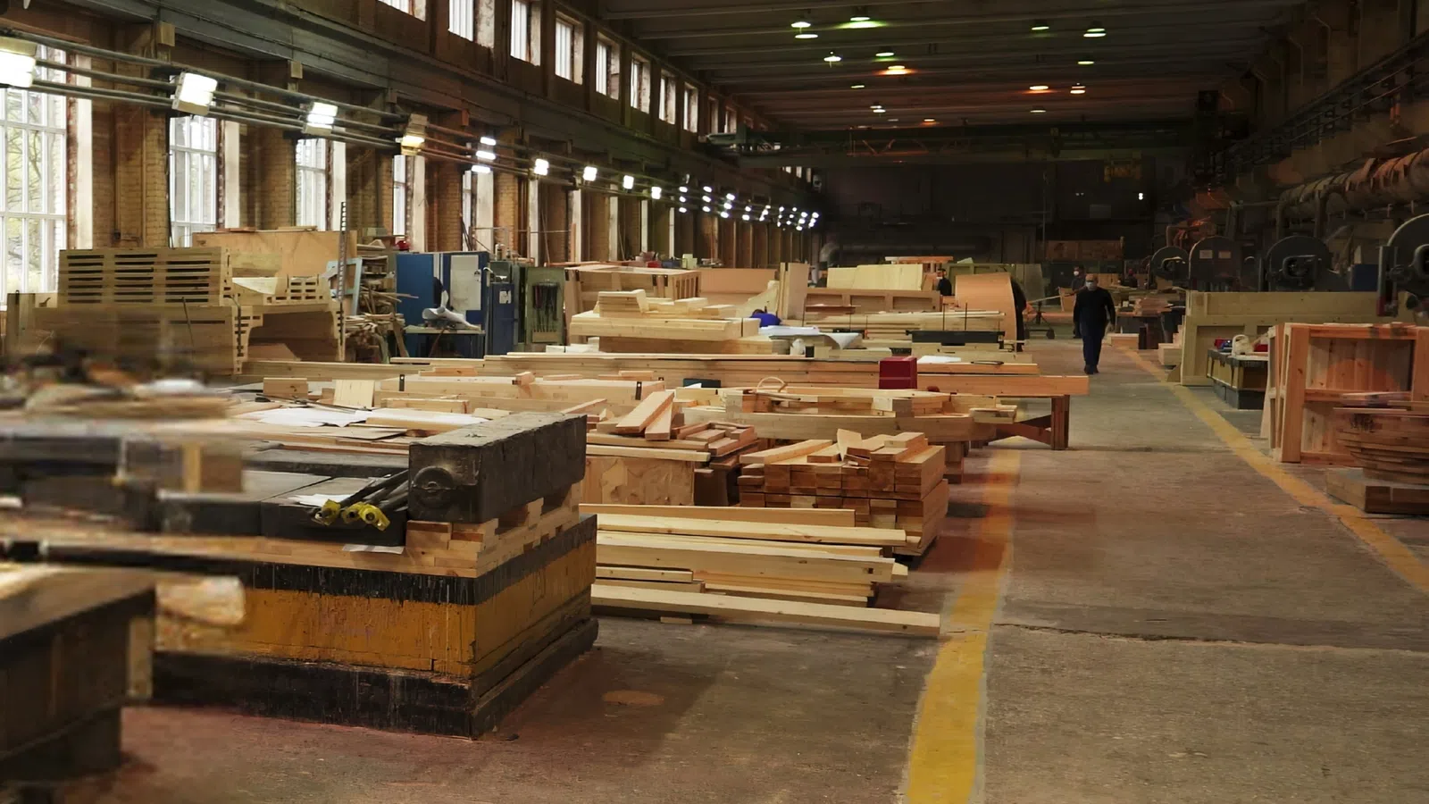 Large woodworking factory interior with stacks of lumber, machinery, and a worker walking down the aisle.