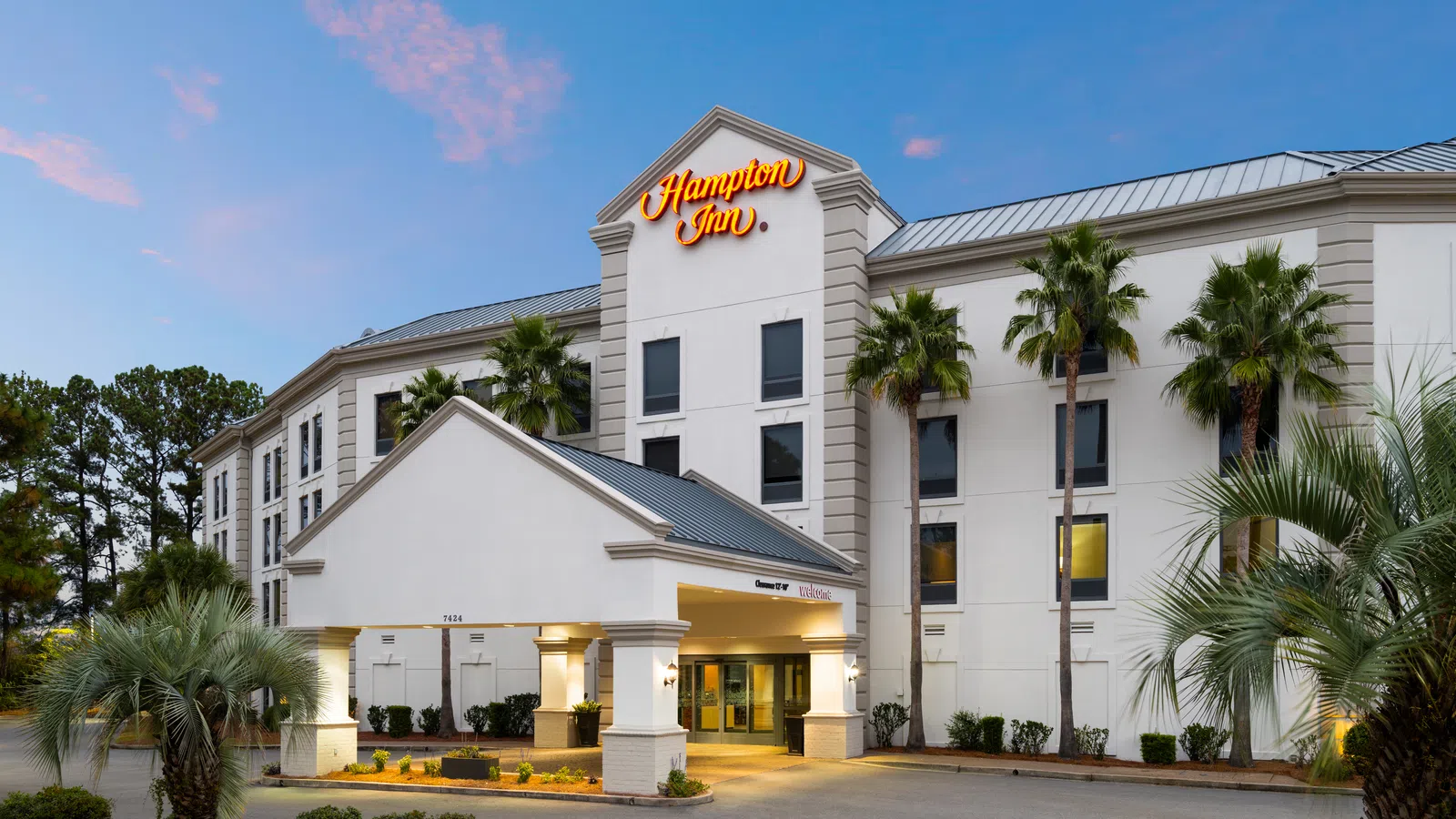 Hampton Inn hotel exterior with palm trees and illuminated sign during early evening light.
