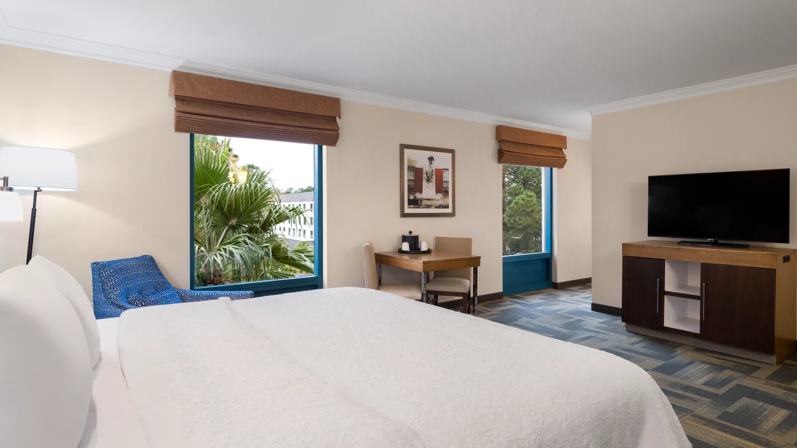 Bright hotel room with a king bed, blue armchair, desk, and TV near windows with brown shades.
