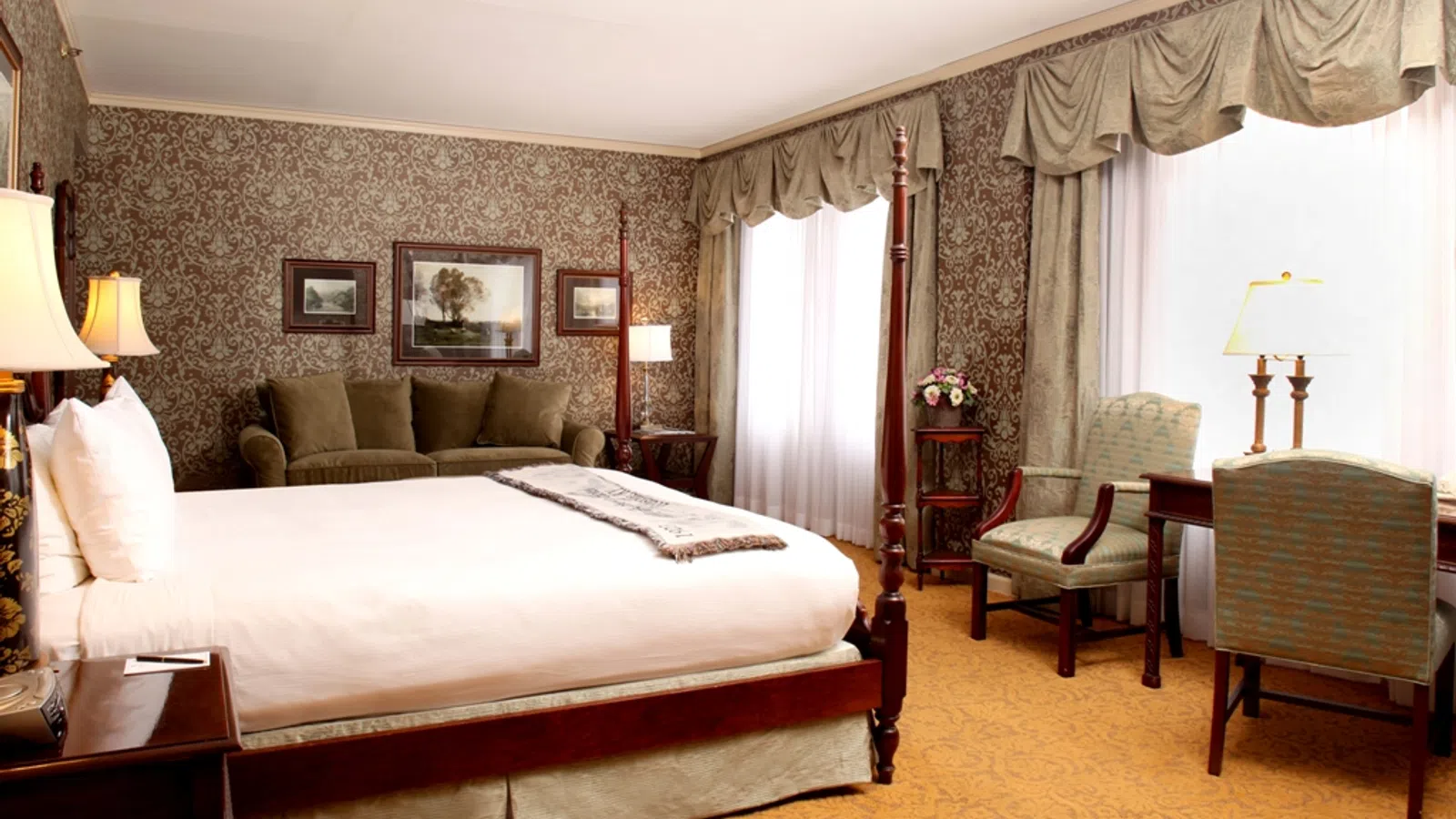 Elegant hotel room with four-poster bed, patterned wallpaper, sofa, desk with chairs, and large windows with curtains.