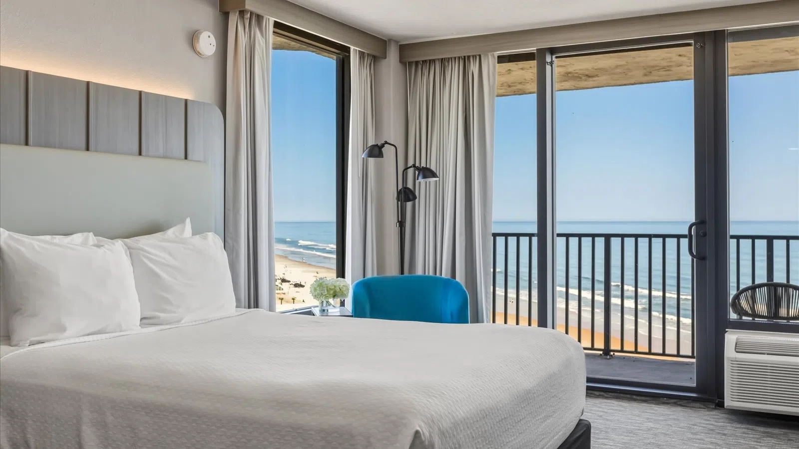 Modern hotel room with king bed, blue chair, ocean view, sliding balcony doors, and natural light.