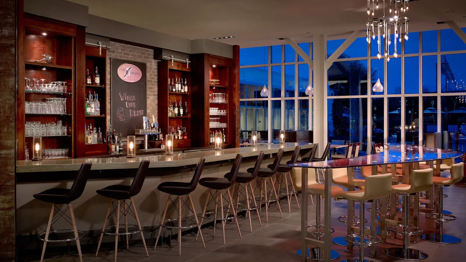 Modern bar with high stools, wooden shelves with bottles, large windows, and elegant lighting in the evening.