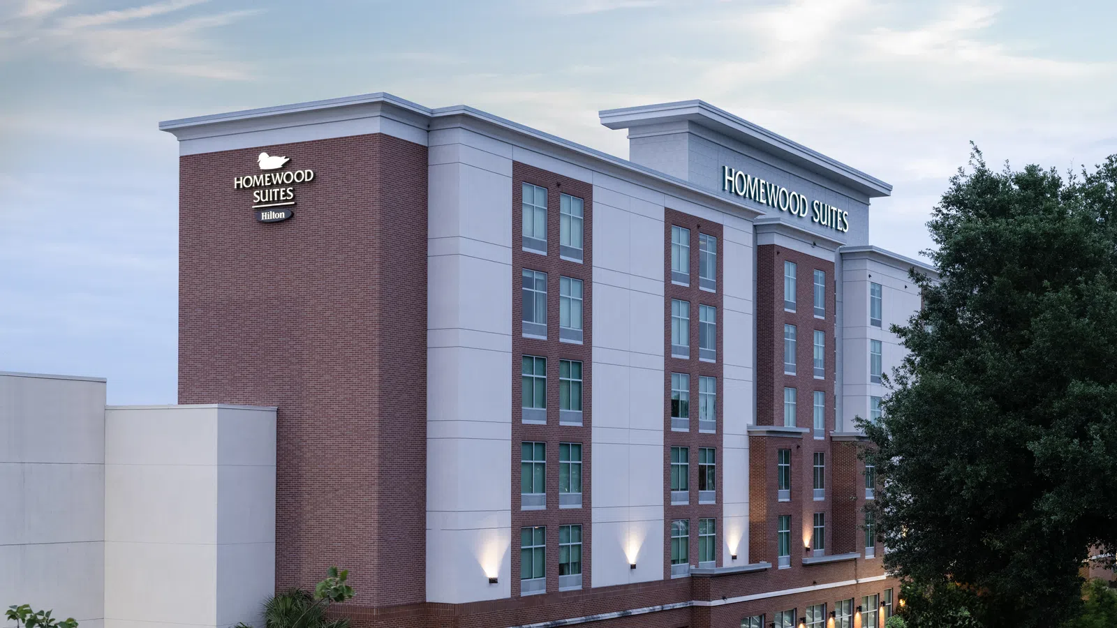 Exterior view of a modern Homewood Suites by Hilton hotel building with trees and evening lighting.