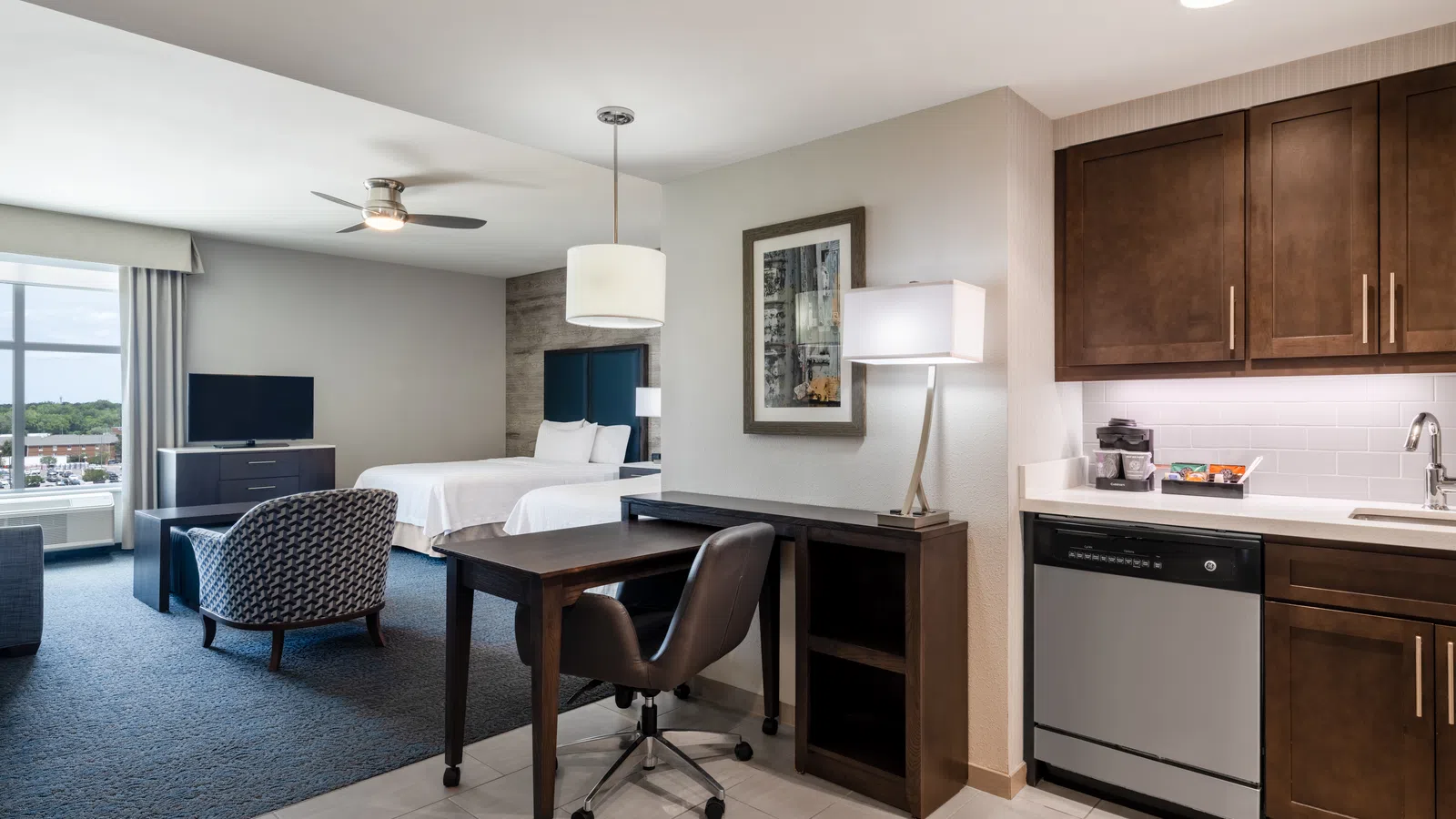 Modern hotel suite with kitchenette, desk, chair, bed, TV, and large window with city view under soft lighting.