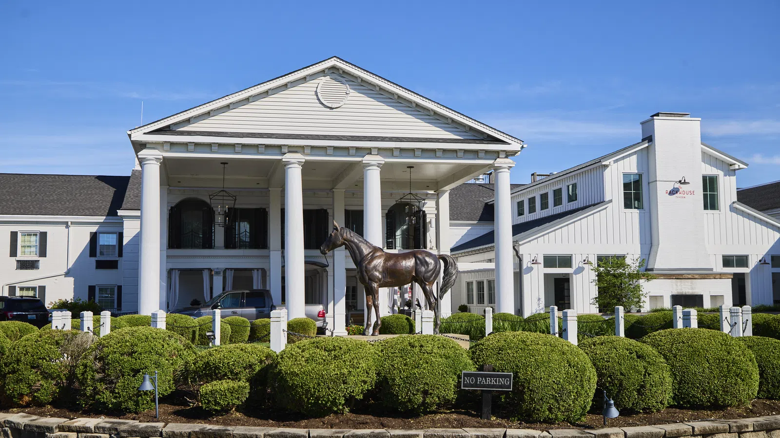 Colonial-style white building with large columns and a bronze horse statue in landscaped circular driveway under clear blue sky
