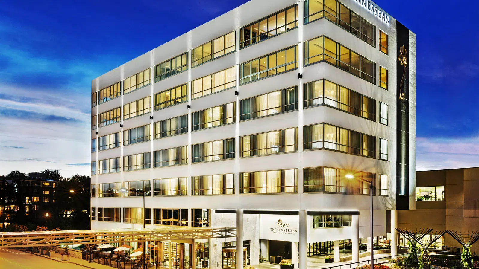 The Tennesseean hotel building illuminated at dusk with large windows and outdoor seating area.